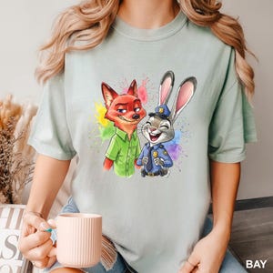 May include: A light green t-shirt featuring a colorful illustration of Nick Wilde the fox and Judy Hopps the rabbit from Zootopia. Nick is wearing a green shirt, and Judy is in a police uniform. The design includes colorful paint splatters.