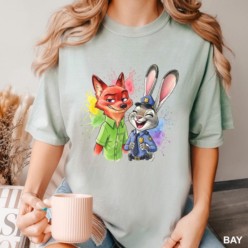 May include: A light green t-shirt featuring a colorful illustration of Nick Wilde the fox and Judy Hopps the rabbit from Zootopia. Nick is wearing a green shirt, and Judy is in a police uniform. The design includes colorful paint splatters.