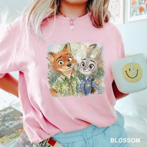 May include: Pink t-shirt featuring a graphic of Nick Wilde and Judy Hopps from Zootopia. The characters are depicted in a colorful, cartoon style. A white mug with a yellow smiley face is held in the hand. The word "BLOSSOM" is printed in white.