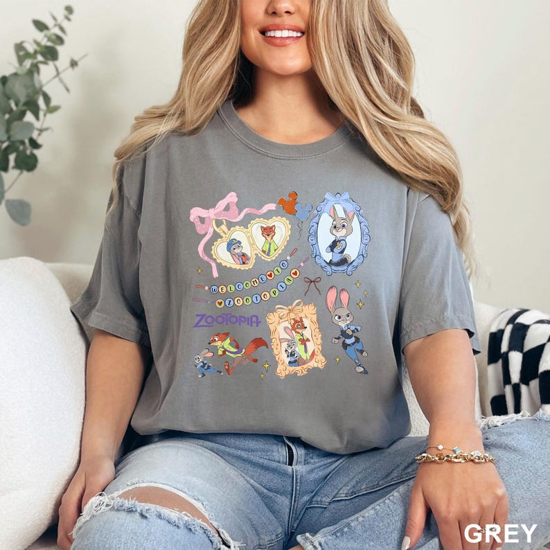 Op de afbeelding: Grijs T-shirt met kleurrijke illustraties van personages uit de animatiefilm "Zootopia". Het ontwerp bevat ingelijste portretten van Judy Hopps en Nick Wilde, samen met de titel van de film. Het shirt is gemaakt van een zacht materiaal.
