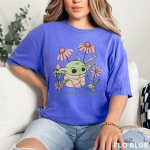 Puede incluir: Camiseta azul con un gr&aacute;fico de un personaje alien&iacute;gena verde rodeado de flores rosas y beige. El texto "Enjoy the little things" est&aacute; impreso debajo. La camiseta es de color azul liso.