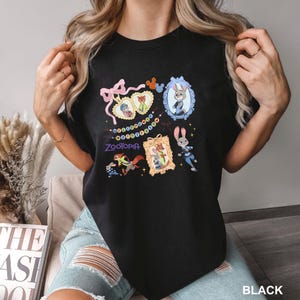 Op de afbeelding: Zwart T-shirt met ronde hals en kleurrijke cartoon illustraties uit de film "Zootopia". Het ontwerp bevat personages in sierlijke frames, een reeks kleurrijke stippen en het logo van de film. Het woord "BLACK" staat rechtsonder gedrukt.