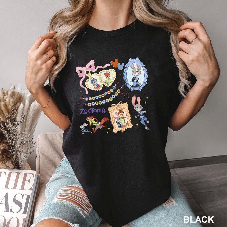 Op de afbeelding: Zwart T-shirt met ronde hals en kleurrijke cartoon illustraties uit de film "Zootopia". Het ontwerp bevat personages in sierlijke frames, een reeks kleurrijke stippen en het logo van de film. Het woord "BLACK" staat rechtsonder gedrukt.
