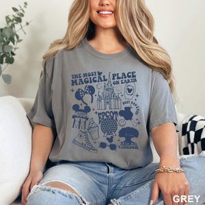 Puede incluir: Camiseta gris con un dise&ntilde;o gr&aacute;fico azul que presenta el texto "THE MOST MAGICAL PLACE ON EARTH" y varios iconos tem&aacute;ticos de Disney. El dise&ntilde;o incluye un castillo, orejas de Mickey Mouse y otras im&aacute;genes relacionadas con el parque.