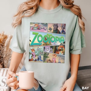 K&ouml;nnte beinhalten: Ein helles salbeigr&uuml;nes T-Shirt mit einem Grafikdruck der Zootopia-Figuren in einem Comic-Panel-Layout. Das Design zeigt verschiedene Szenen mit Nick Wilde und Judy Hopps, wobei das Wort Zootopia in gro&szlig;en gr&uuml;nen Buchstaben gedruckt ist.