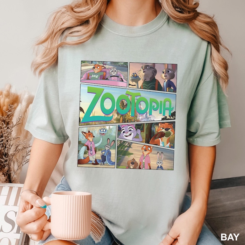K&ouml;nnte beinhalten: Ein helles salbeigr&uuml;nes T-Shirt mit einem Grafikdruck der Zootopia-Figuren in einem Comic-Panel-Layout. Das Design zeigt verschiedene Szenen mit Nick Wilde und Judy Hopps, wobei das Wort Zootopia in gro&szlig;en gr&uuml;nen Buchstaben gedruckt ist.