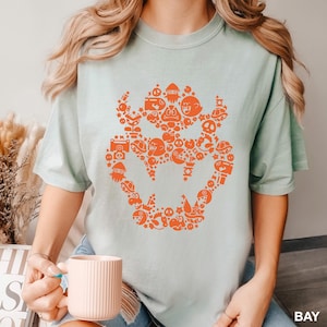 May include: A light green t-shirt featuring a large orange graphic of a character's face composed of various cartoon elements. The shirt has short sleeves and a relaxed fit. The word "BAY" is printed in white in the bottom right corner.