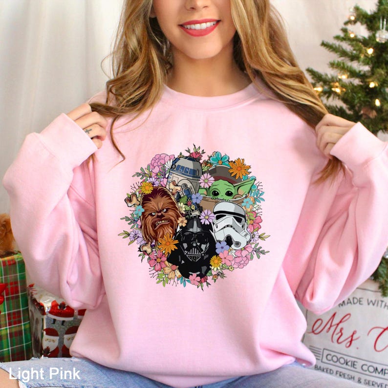 Op de afbeelding: Lichtroze sweatshirt met een bloemenontwerp met Star Wars-personages zoals Darth Vader, Chewbacca en Baby Yoda. Het ontwerp is omgeven door kleurrijke bloemen. De sweatshirt heeft een ronde hals.
