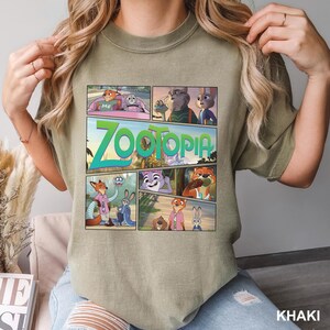 K&ouml;nnte beinhalten: Ein khakifarbenes T-Shirt mit Rundhalsausschnitt und einem gro&szlig;en Grafikdruck verschiedener Szenen aus dem Film Zoomania. Das Design enth&auml;lt mehrere Felder mit animierten Charakteren wie Nick Wilde und Judy Hopps sowie dem Wort Zootopia in Gr&uuml;n.