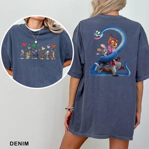 Op de afbeelding: Denim blauw T-shirt met cartoonfiguren op de voor- en achterkant. De voorkant heeft een rij karakters met kleurrijke vormen erboven. De achterkant toont een vos, konijn en slang ontwerp.