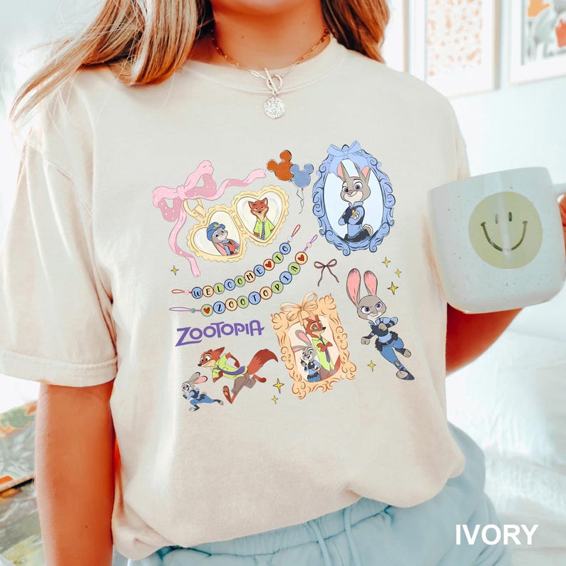 Op de afbeelding: Ivoor T-shirt met kleurrijke Zootopia-karakterillustraties. Het ontwerp bevat ingelijste portretten van Judy Hopps en Nick Wilde, het woord "Zootopia" en de zin "Welcome to Zootopia". Een witte mok met een smiley is ook zichtbaar.