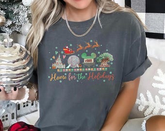 May include: A charcoal gray short-sleeve t-shirt featuring a graphic print of a Santa sleigh, reindeer, and four theme park landmarks. The text "Home for the Holidays" is printed in a cursive font below the icons, accented by small snowflakes.