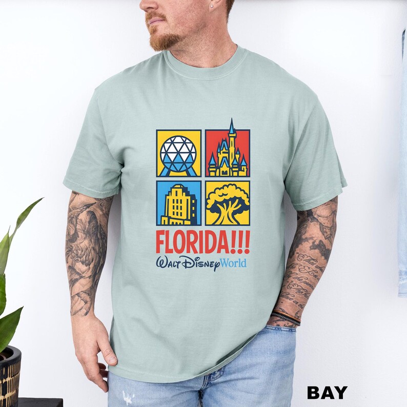 Może przedstawiać: Jasnozielona koszulka z kolorową grafiką przedstawiającą zabytki Florydy. Projekt zawiera tekst "FLORIDA!!!" i "Walt Disney World". Koszulka ma kr&oacute;tkie rękawy i okrągły dekolt.