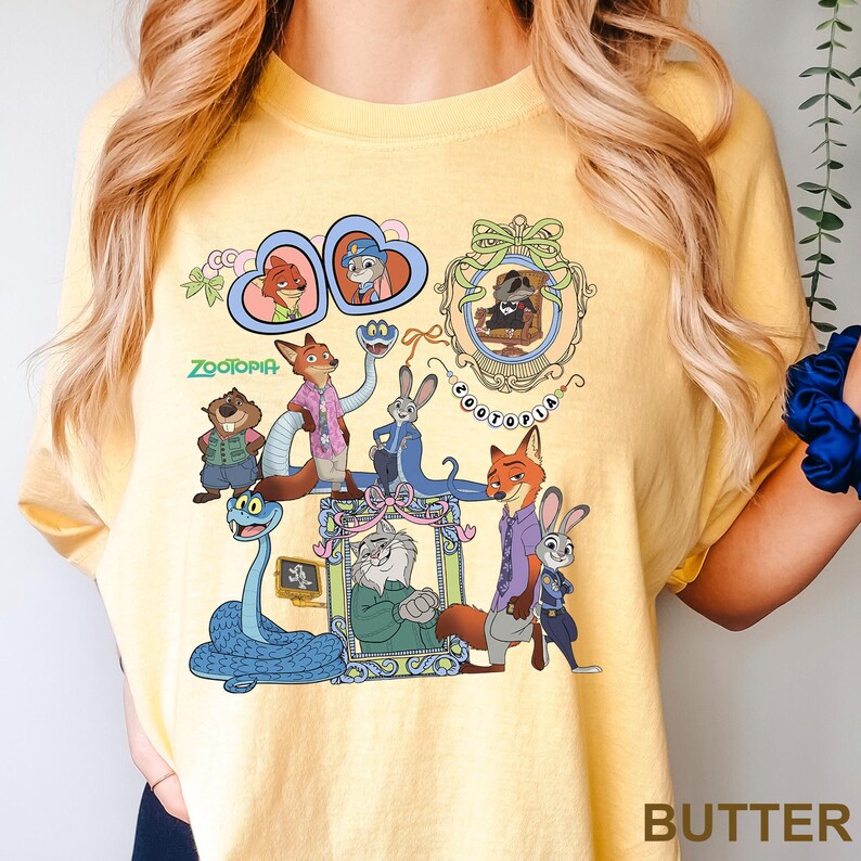 May include: A butter yellow t-shirt featuring a colorful graphic print of Zootopia characters including Nick Wilde, Judy Hopps, and others. The design includes various framed portraits, a blue snake, and the Zootopia logo text in green and white.