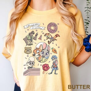 Op de afbeelding: Geel T-shirt met cartoonfiguren uit Zootopia. Het ontwerp bevat Nick Wilde, Judy Hopps en andere personages, samen met de tekst "You can be whatever you wish to be" en "The Big Donut".