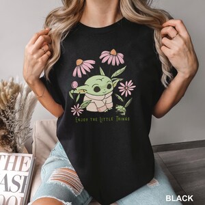 Puede incluir: Camiseta negra con un personaje de dibujos animados verde, flores rosas y hojas verdes. El texto "ENJOY THE LITTLE THINGS" est&aacute; impreso en verde. La palabra "BLACK" est&aacute; impresa en la parte inferior.