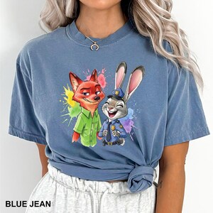 May include: Blue jean colored t-shirt featuring a colorful graphic of the Zootopia characters Nick Wilde and Judy Hopps. Nick is a red fox wearing a green shirt, and Judy is a gray rabbit in a police uniform. The text "BLUE JEAN" is at the bottom.