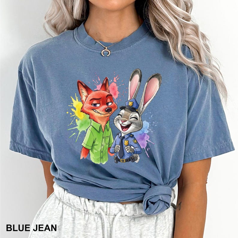 May include: Blue jean colored t-shirt featuring a colorful graphic of the Zootopia characters Nick Wilde and Judy Hopps. Nick is a red fox wearing a green shirt, and Judy is a gray rabbit in a police uniform. The text "BLUE JEAN" is at the bottom.