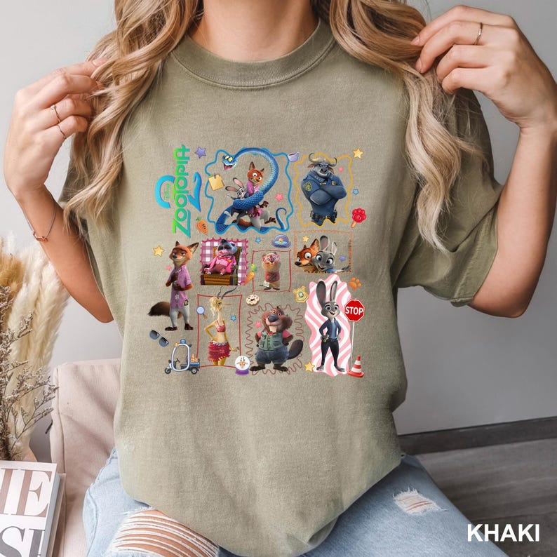 May include: A khaki-colored crew neck t-shirt featuring a colorful graphic print of various animated characters from the movie Zootopia. The design includes multiple framed scenes, small icons, and the word Zootopia written vertically in bright green text.