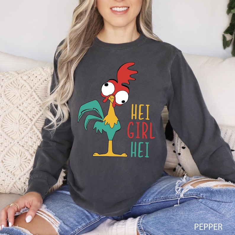 May include: Dark gray long-sleeve sweatshirt featuring a cartoon rooster with red, teal, yellow, and white accents. The text "HEI GIRL HEI" is printed in teal, red, and yellow. The sweatshirt is made of a soft material.