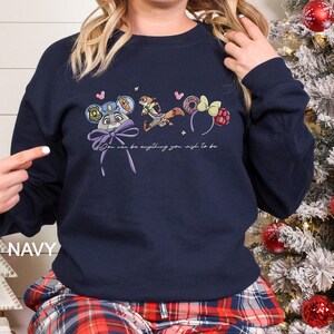 May include: Navy blue sweatshirt with a cartoon design of a bunny, fox, and themed headbands, along with the text "You can be anything you wish to be." The design includes a purple bow. The sweatshirt is a dark blue colour.