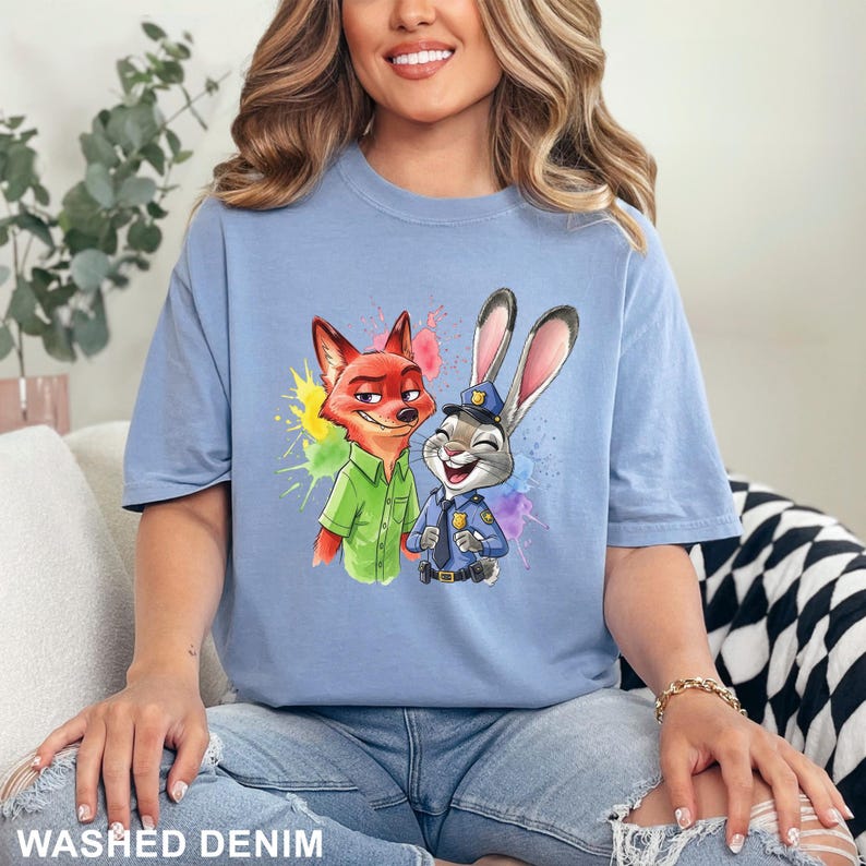 May include: Light blue t-shirt featuring a watercolor illustration of Nick Wilde and Judy Hopps from Zootopia. The design includes colorful paint splatters. The words "Washed Denim" are printed at the bottom of the image.