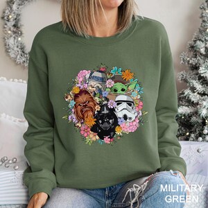Op de afbeelding: Militairgroen sweatshirt met een bloemenontwerp met Star Wars-personages zoals Darth Vader en Baby Yoda. De sweatshirt heeft een ronde hals en lange mouwen. De tekst "MILITARY GREEN" is zichtbaar.