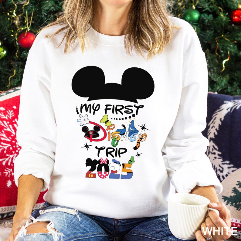 Puede incluir: Sudadera blanca con una silueta negra de Mickey Mouse y texto colorido "My First Disney Trip 2025". El dise&ntilde;o incluye a Mickey Mouse, un gorro de chef, una mariposa y un tigre.