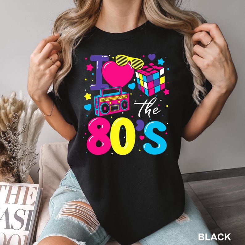 K&ouml;nnte beinhalten: Schwarzes T-Shirt mit einem farbenfrohen Aufdruck "I love the 80s". Das Design umfasst ein pinkes Herz, gelbe Sonnenbrillen, einen Ghettoblaster, einen Rubik's Cube und Sterne. Das Wort "BLACK" ist unten rechts aufgedruckt.