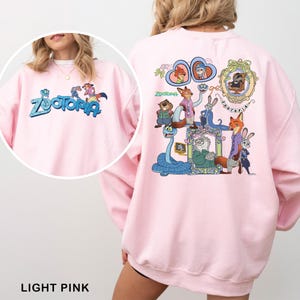 Puede incluir: Sudadera rosa claro con la palabra "Zootopia" en azul y personajes de dibujos animados en la parte delantera y trasera. El dise&ntilde;o trasero incluye im&aacute;genes enmarcadas de personajes y la palabra "Zootopia".