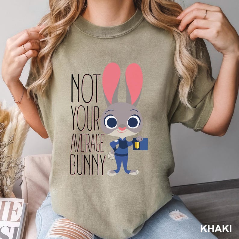 May include: Khaki-colored t-shirt featuring a cartoon bunny in a police uniform, with the text "NOT YOUR AVERAGE BUNNY" in black. The bunny has pink ears and is holding a briefcase. The shirt is a relaxed fit.