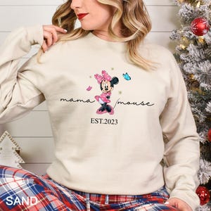 May include: A cream-colored sweatshirt with a graphic of Minnie Mouse in a pink dress and red shoes. The text "mama mouse EST.2023" is printed below the graphic.