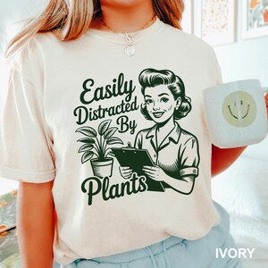 May include: An ivory short-sleeve t-shirt featuring a dark green graphic print of a person holding a clipboard next to a potted plant. The text reads Easily Distracted By Plants. The shirt is paired with a white mug with a yellow smiley face.