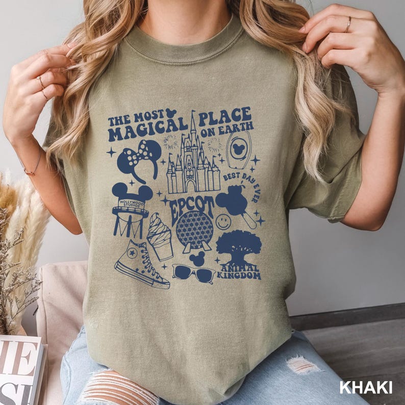 Puede incluir: Camiseta caqui con un dise&ntilde;o azul marino que dice "The Most Magical Place on Earth". El dise&ntilde;o incluye iconos de un castillo, orejas de Mickey Mouse y un helado. Una prenda de vestir informal.