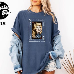 May include: A denim blue short-sleeve t-shirt featuring a graphic print of a playing card with a portrait of a blonde woman and the name Dolly written in script. The card design includes the letter Q and heart symbols in the corners.