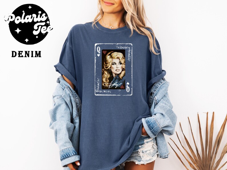 May include: A denim blue short-sleeve t-shirt featuring a graphic print of a playing card with a portrait of a blonde woman and the name Dolly written in script. The card design includes the letter Q and heart symbols in the corners.