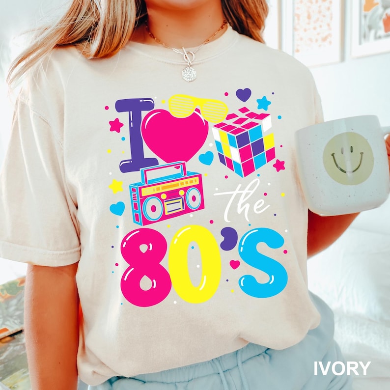K&ouml;nnte beinhalten: Elfenbeinfarbenes T-Shirt mit einem farbenfrohen 80er-Jahre-Motiv. Das Design umfasst ein pinkes Herz, eine Sonnenbrille, einen Zauberw&uuml;rfel, einen Ghettoblaster und den Text "I love the 80's". Eine wei&szlig;e Tasse mit einem Smiley wird im Bild gehalten.