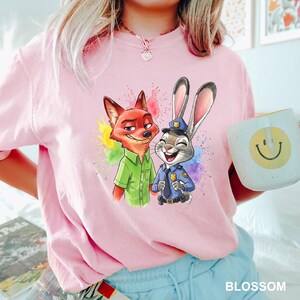 May include: Pink t-shirt featuring a colorful graphic of Nick Wilde and Judy Hopps from Zootopia, with paint splatter accents. The shirt also has the word "BLOSSOM" printed at the bottom. A mug with a smiley face is held in the image.