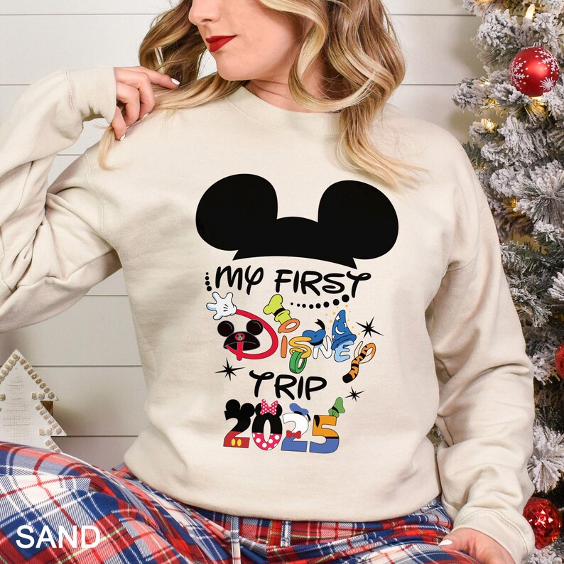 Puede incluir: Una sudadera de color arena con una silueta negra de Mickey Mouse y el texto "My First Disney Trip 2025" en letras coloridas. La sudadera tiene mangas largas y cuello redondo.