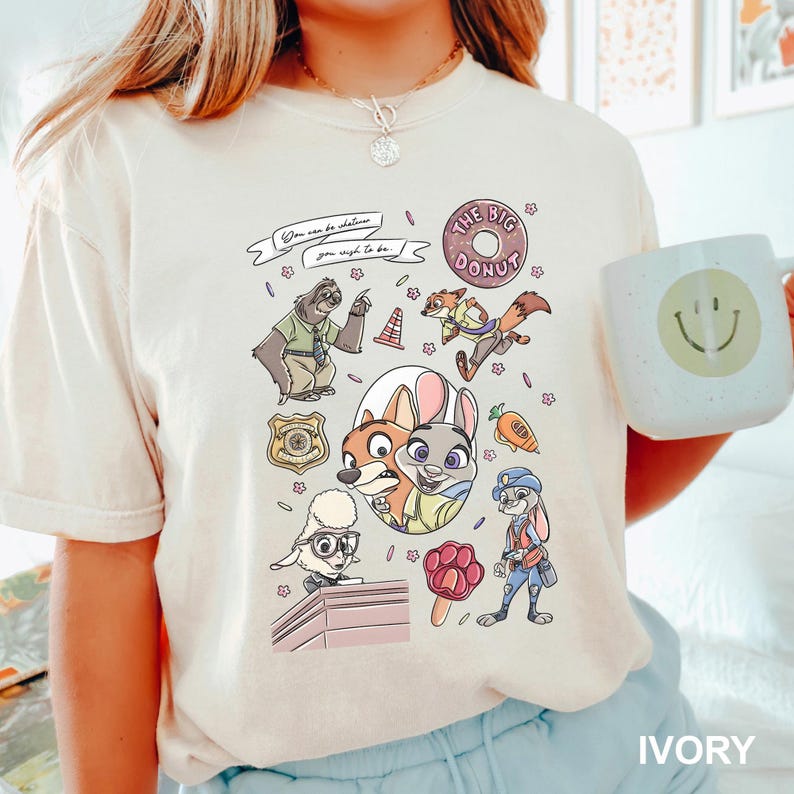 Op de afbeelding: Ivoor t-shirt met een kleurrijke cartoonafbeelding van personages. Het ontwerp bevat een donut, een politiebadge en de tekst "You can be whatever you wish to be."