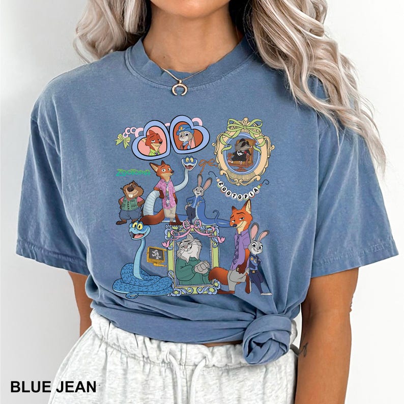 May include: Blue jean coloured t-shirt with a colourful graphic of cartoon animals and the word "Zootopia". The graphic includes a fox, rabbit, and other characters in a whimsical design. The shirt has a crew neck and short sleeves.