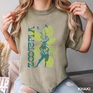 May include: Khaki-colored t-shirt featuring a graphic of Judy Hopps from Zootopia. The design includes the word "Zootopia" in teal, along with a cartoon image of Judy Hopps in a police uniform, surrounded by green tropical leaves.