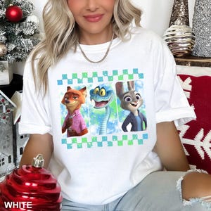 May include: White t-shirt featuring a colorful graphic of three cartoon characters: a fox, a snake, and a rabbit. The design is framed by a green and blue checkered pattern. The word "WHITE" is printed at the bottom.