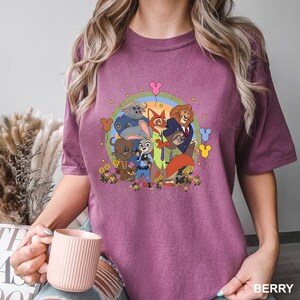 Pode incluir: Camiseta cor de berry com um gr&aacute;fico colorido de personagens de Zootopia. O design inclui Judy Hopps, Nick Wilde e outros personagens em um arranjo circular com flores e bal&otilde;es em forma de Mickey Mouse. A camisa &eacute; um estilo casual e confort&aacute;vel.