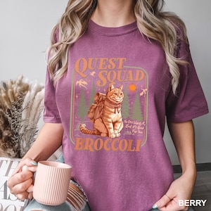 May include: Berry-colored t-shirt featuring a graphic of a cat with a backpack, the words "Quest Squad" and "Broccoli". The cat is in a forest scene with trees and a sun. The text "No One Wants It, But It's Good For You" is also included.
