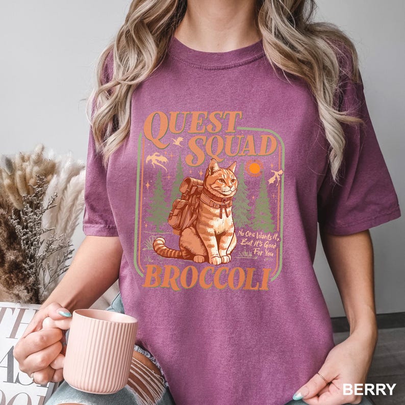 May include: Berry-colored t-shirt featuring a graphic of a cat with a backpack, the words "Quest Squad" and "Broccoli". The cat is in a forest scene with trees and a sun. The text "No One Wants It, But It's Good For You" is also included.