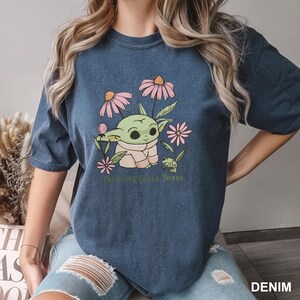 Puede incluir: Camiseta azul denim con un gr&aacute;fico de un personaje verde alien&iacute;gena rodeado de flores rosas y el texto "Enjoy the Little Things". La camiseta es de corte holgado.