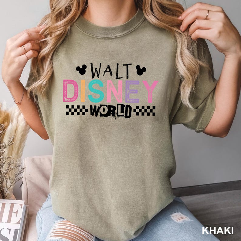 May include: A khaki-colored short-sleeve t-shirt featuring the text WALT DISNEY WORLD in a multi-colored, distressed font. The design includes black Mickey Mouse silhouettes and a black and white checkered pattern below the word WORLD.