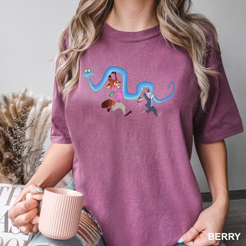 Op de afbeelding: Bessenkleurig T-shirt met een geborduurd ontwerp van een vos en een konijn die rennen met een blauwe slang. Het shirt heeft korte mouwen en een ronde hals. Het woord "BERRY" staat rechtsonder gedrukt.