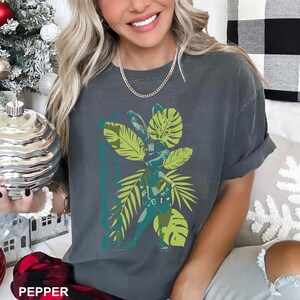 May include: Dark grey t-shirt with a graphic of a figure surrounded by green and yellow tropical leaves. The word "PEPPER" is at the bottom of the shirt. The figure is wearing a belt and has bunny ears.